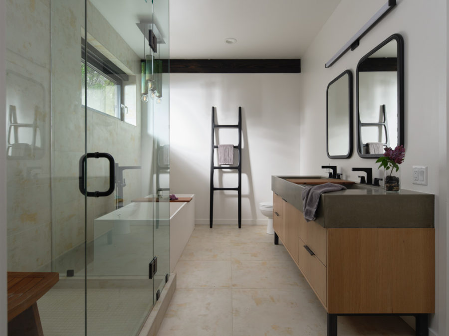 Modern bathroom with double vanity, stone floors, large glass-wall shower. Joists in ceiling are visible. Dark wood.